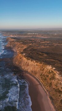 Drone flying along cliffs near Ribat da Arrifana in Algarve, Portugal. Golden sunset light reveals wild Atlantic coastline and empty beach below