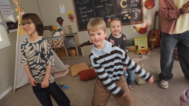 Handheld shot of joyful Caucasian girls and boys dancing together with young biracial male educator, jumping and having fun during physical activity in cozy kindergarten classroom