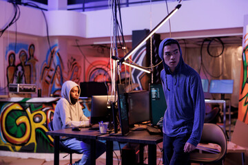 Fototapeta premium Suspicious hacker looking away after breaking into server and doing illegal activity. Young asian criminals in hood leaving abandoned warehouse after breaching database and stealing data