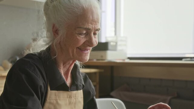Low angle tilt up shot of elderly craftswoman with gray hair smiling and peacefully painting heart on flat peice of clay with brush in modern pottery studio