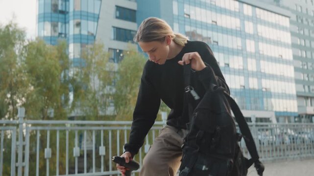 Caucasian urban explorer unpacking gear near modern waterfront, walking between glass buildings, kneeling to open backpack, methodical gear check, handheld tool and charger visible, daytime cityscape