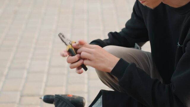 Young man checking small tool and battery. Male kneeling on pavement inspecting tool with screwdriver. Young man crouching on city sidewalk carefully inspecting equipment with tools and battery