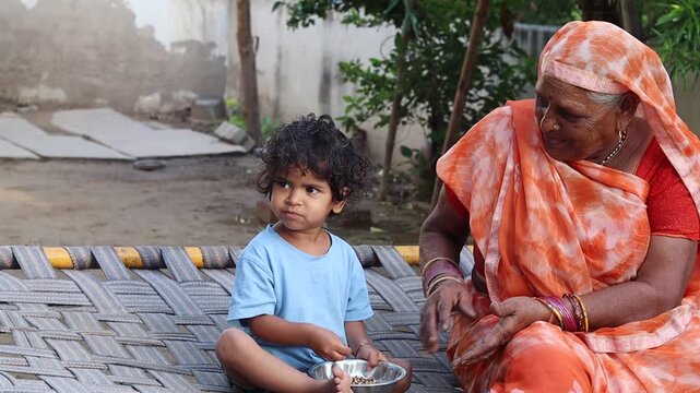 An affectionate elderly Indian grandmother sitting on a traditional woven cot (charpai) outdoors, lovingly watching her toddler grandchild eat from a small stainless steel bowl, highlighting rural fam