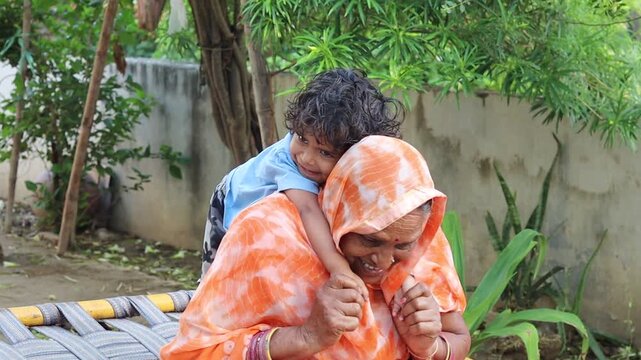 Candid shot of a loving Indian grandmother carrying a toddler on her back, sitting on a charpai in a rural courtyard.