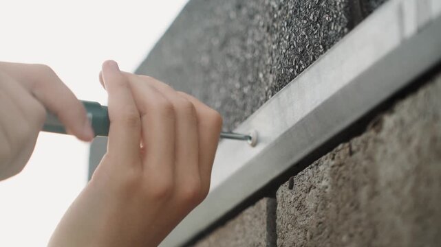 Hands tightening screw along flashing, brick edge under neutral daylight, twohand grip turning compact screwdriver, steady motion, routine exterior maintenance, ensuring secure attachment
