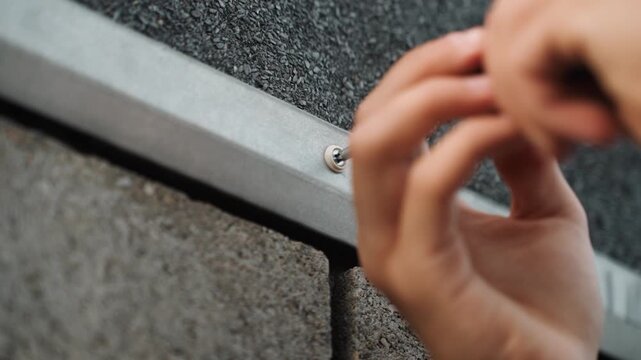 Closeup hands tightening roof flashing screw, handyman using screwdriver to secure metal flashing under asphalt shingle on exterior wall, concrete edge visible, focused shallow depth of field,