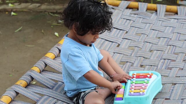 A cute little Indian toddler girl sitting on a traditional woven cot (charpai) outdoors, playfully engaging with a colorful musical toy piano, representing early childhood learning and development.
