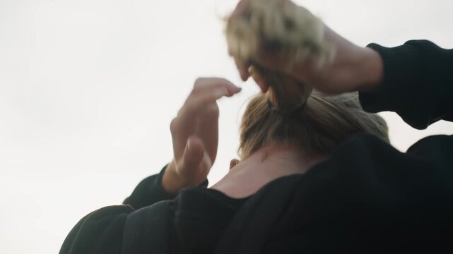 Blonde hands tying ponytail outdoors before run focused gesture secures elastic, wind and daylight, athletic mood, commuter or runner preparing, close framing on back of head and neck, tactile detail