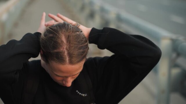 White teen male smiling tying braid energetic styling cornrows on pedestrian bridge golden hour smile hands styling hair casual black sweatshirt urban railing background candid lifestyle shot cheerful