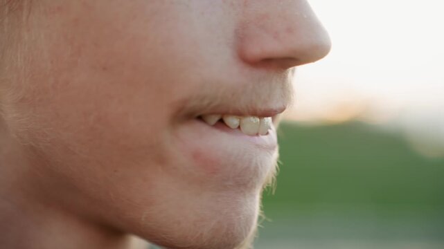 White young man smiling playfully at sunset, lip movement and light tongue gesture, freckled chin with light stubble, closeup mouth and jaw, relaxed friendly mood, warm natural light, candid outdoor