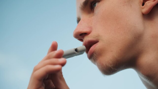 Lowangle caucasian traveler vaping under blue sky, youthful male inhaling from compact mouthpiece, windtousled hair, carefree summer vibe, outdoor pause between activities, sunlight on jawline, brisk
