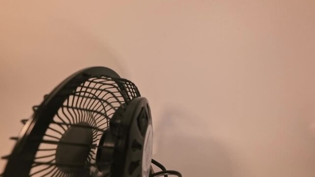 A hand reaches out to press the power button on a black desk fan, instantly activating the blades to provide a cool breeze against a plain wall.