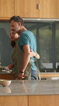 Vertical video: Hug releasing, couple moving to stove at kitchen island in aprons, stirring pan