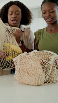 Vertical video: Teen in olive top placing mesh bag in kitchen, untangling and unpacking groceries