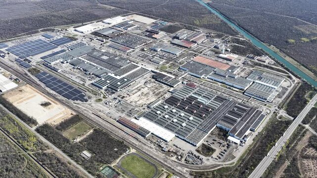 Aerial view of a sprawling industrial complex featuring many buildings and large solar panel arrays, framed by lush greenery, Sausheim, Grand Est, France.