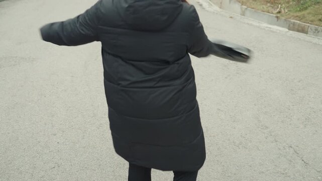 Woman in black coat dancing playfully on empty road, twirling tote bag while wearing gray beanie, smiling and waving, visible white arrow on pavement, cool winter light, urban commuter choreography