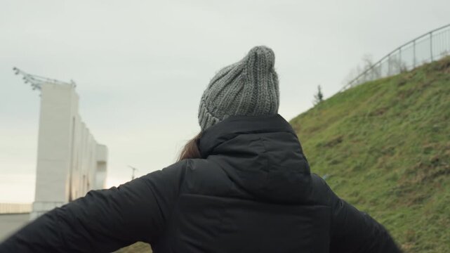 Open arm winter dance on promenade, woman with grey beanie and puffer jacket faces skyline with arms outstretched, overcast sky and railing backdrop, slow expressive movement and reflective mood