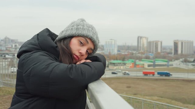 Contemplative rooftop dance with white woman leaning on railing, gray beanie and black puffer coat against overcast city skyline, slow expressive choreography, closeup gaze and reflective mood, subtle