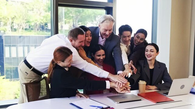 A diverse office team gathers around a desk, stacking hands in a spirited huddle then raise, smiling and energized during a meeting with laptops and documents nearby, symbolizing teamwork and unity.