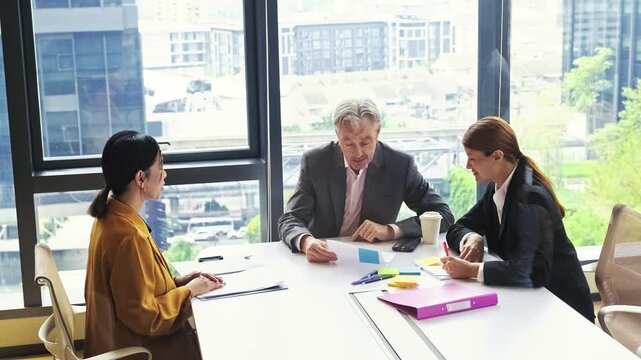 Three professionals in a glass-walled office hold a meeting at a conference table, reviewing documents and notes with a city skyline visible through large windows.