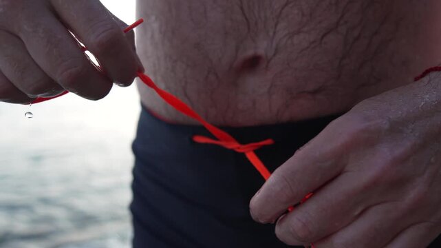 Swimmer tying orange drawstring on dark swimsuit bottoms at a rocky beach shore before swimming in the ocean