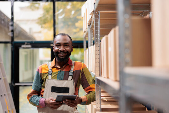 Portrait of smiling employee working storage room, preparing customers packages for delivery. Storage room worker supervising cardboard boxes in mail sorting center, writing inventory on clipboard