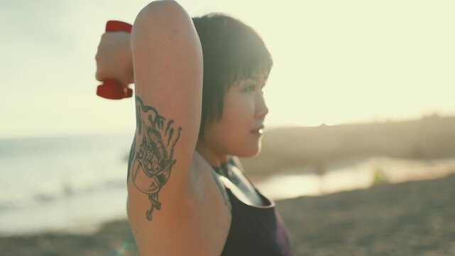 Tattooed woman training triceps with dumbbell on the beach