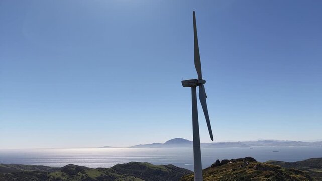 Close-up back tracking drone shot of a wind turbine against a clear blue sky with the Strait of Gibraltar and Africa in the distance.