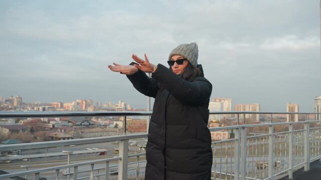 City skyline dance with panoramic view woman in warm coat and gray beanie moves through measured gestures on rooftop balcony, distant city blocks, cinematic golden hour light, adjusts sunglasses,