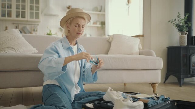 Stylish elderly woman packing suitcase for summer trip, putting clothes and map. Happy female traveler dreaming about tourism and traveling in unknown city, traditional paper map for navigation