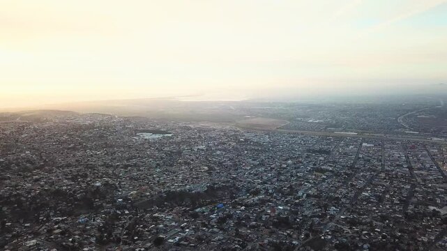 Aerial drone view of Tijuana cityscape toward the coastline, capturing dense urban sprawl under warm sunset light.