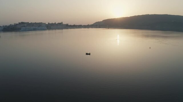 Single fishing boat floats on calm Lake Pichola waters at golden hour sunrise with Aravalli Hills backdrop, Udaipur, Rajasthan. Palace architecture visible along distant shoreline.
