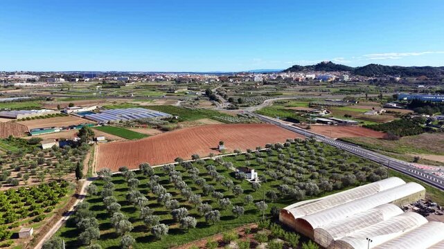 Aerial view of Valencia region farmlands showing plowed red soil fields, olive groves, greenhouses, and a highway stretching toward a hilltop town