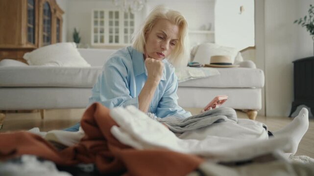 Aged women chatting by video call when packing suitcase for summer traveling. Active lifestyle after 60 years and better aging, happy female traveler putting clothes in travel bag, medium shot