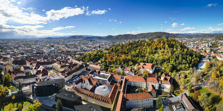 Aerial view of the vibrant red rooftops contrasting with the lush green Schlossberg hill under a bright blue sky, Graz, Styria, Austria.