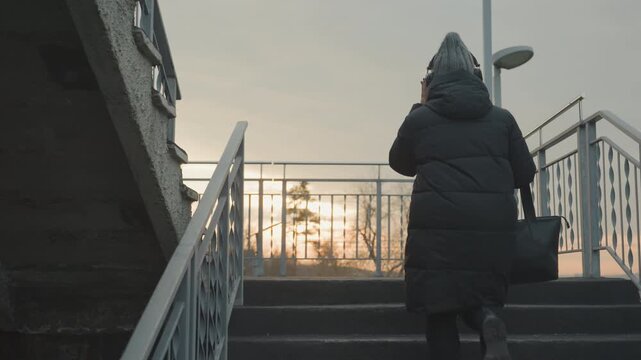 Woman ascending metal stairs at sunrise wearing puffy coat and tote, stepping to internal rhythm and occasional dance sway, city horizon glowing, solitary commuter vibe with calm light