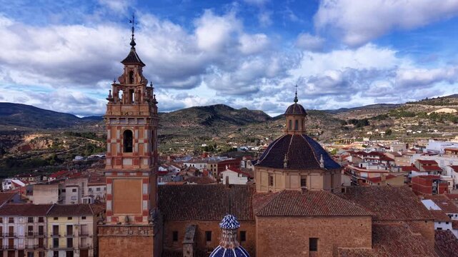 Chelva town in Valencia province, Spain, featuring a baroque church bell tower and blue-tiled dome rising above terracotta rooftops with mountains beyond