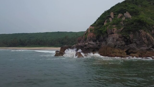 Drone moves forward over turquoise ocean waves approaching rugged brown rock cliffs at Bhogve Beach, Malvan, Maharashtra. Sandy beach and green vegetation visible beyond rocky coastline.