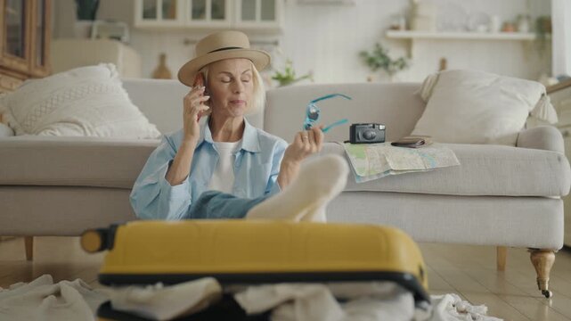 Carefree mature woman relaxing at home after travel, talking by phone, portrait. Traveling and tourism, enjoy summertime and weekend trip, beautiful senior lady sitting on floor near packed suitcase