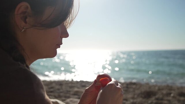 Woman beach fruit enjoying a peaceful moment peeling a fresh nectarine on the pebble shore during a golden sunset