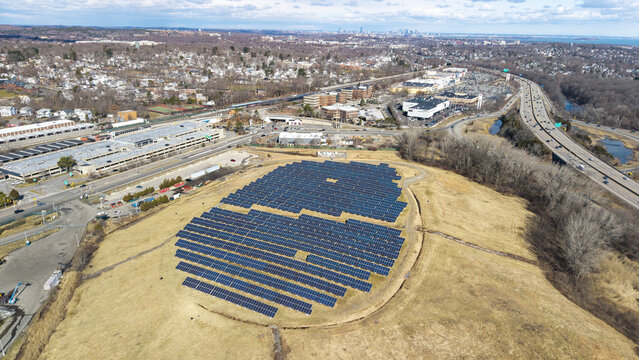 Aerial view of rows of solar panels glinting under the sun on a grassy hill near a highway, contrasting with the urban sprawl, Braintree, Massachusetts, United States.