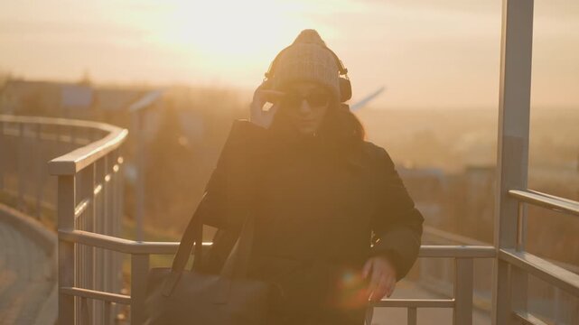 Golden hour dance and reveal. Woman removes sunglasses while moving toward camera, playful glance, sunflare halo, knit hat and winter coat, rhythmic motion and intimate portrait composition.