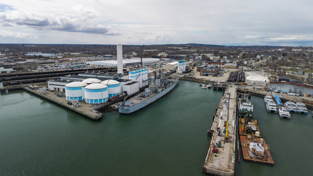Aerial view of the USS Salem resting in the Fore River, surrounded by bright white storage tanks and a working dry dock, Quincy, Massachusetts, United States.