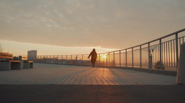 Sunrise promenade silhouette dancer moving across tiled walkway. Lone performer executes fluid contemporary choreography along metal railing as warm golden light casts long shadows over empty urban