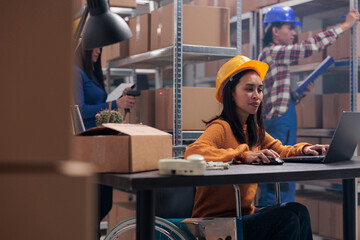 Asian postal service worker with physical disability using laptop in warehouse. Young woman wearing...