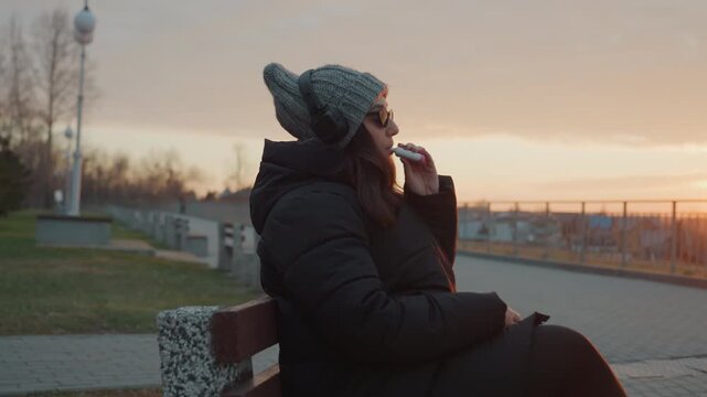 Woman on bench dancing to music at sunset, wearing knit hat and headphones, sunglasses glinting, warm orange rim light, slow hand gestures, relaxed smile, urban park path and skyline in background,