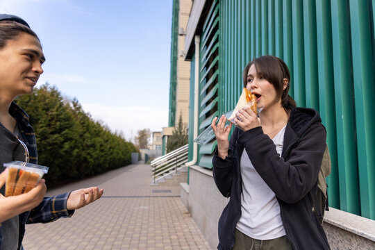 Close up of a young girl with a backpack eating a sandwich from a plastic box while talking to a friend.