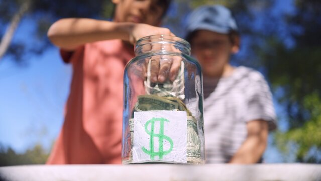 Children learning about saving money and financial literacy. Putting dollar bills into a transparent jar with a hand-drawn money symbol