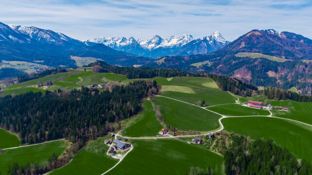 Aerial view of rolling green hills meeting dark forests, crowned by snow-capped mountains under a bright sky, Windischgarsten, Upper Austria, Austria.