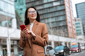 Fototapeta premium Smiling Asian woman businesswoman in tan blazer holding red smartphone for trade business work on city street at office building. Urban professional lifestyle, communication and technology. Copy space